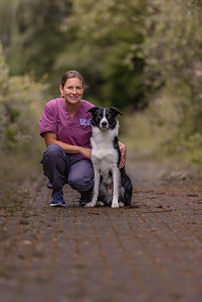 Laechelnde Frau in violetter Kleidung kniet neben einem Border Collie im Wald