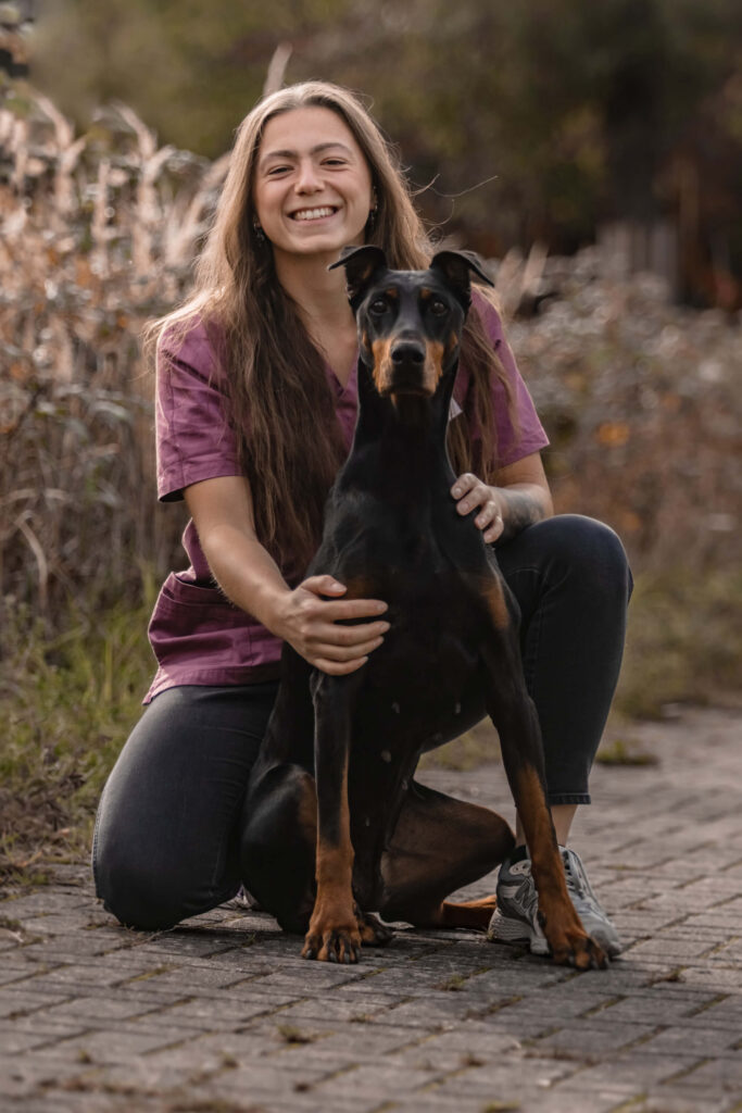 Lachende junge Frau im lila Oberteil kniet mit einem schwarzbraunen Dobermann