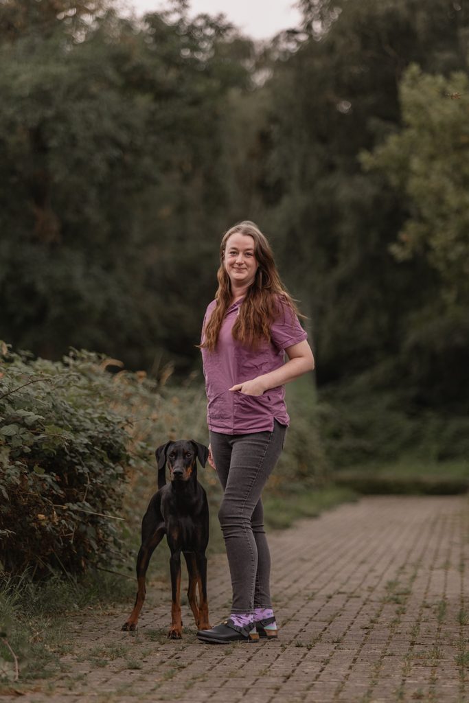 Laechelnde Frau in lila Kittel steht mit schwarzem Dobermann im Gruenen
