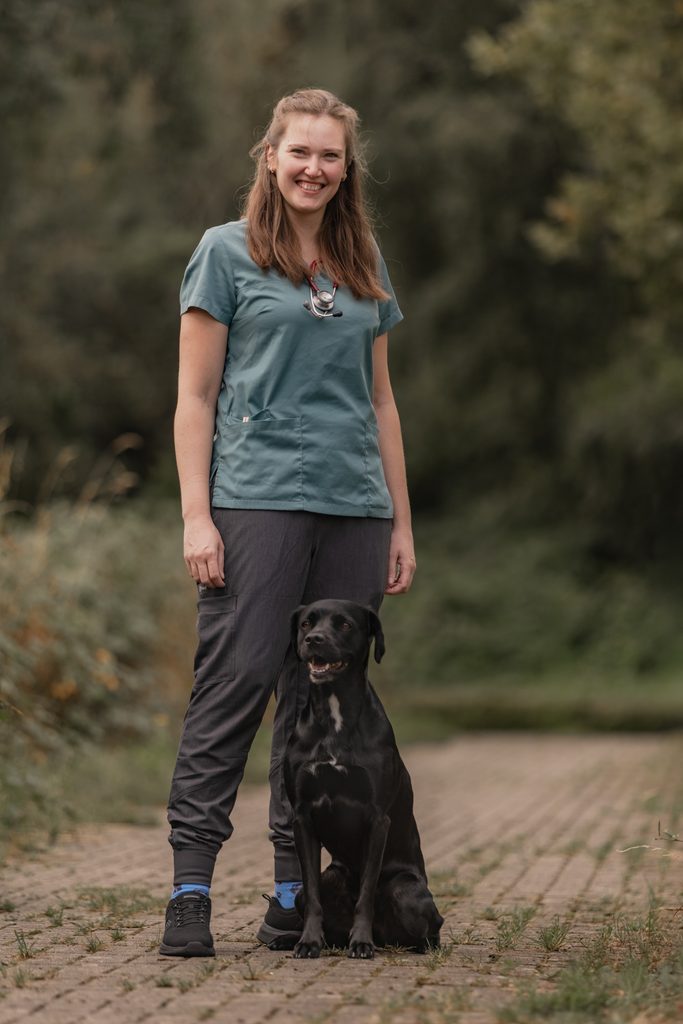 Laechelnde Tieraerztin in gruener Kleidung steht neben schwarzem Hund im Freien